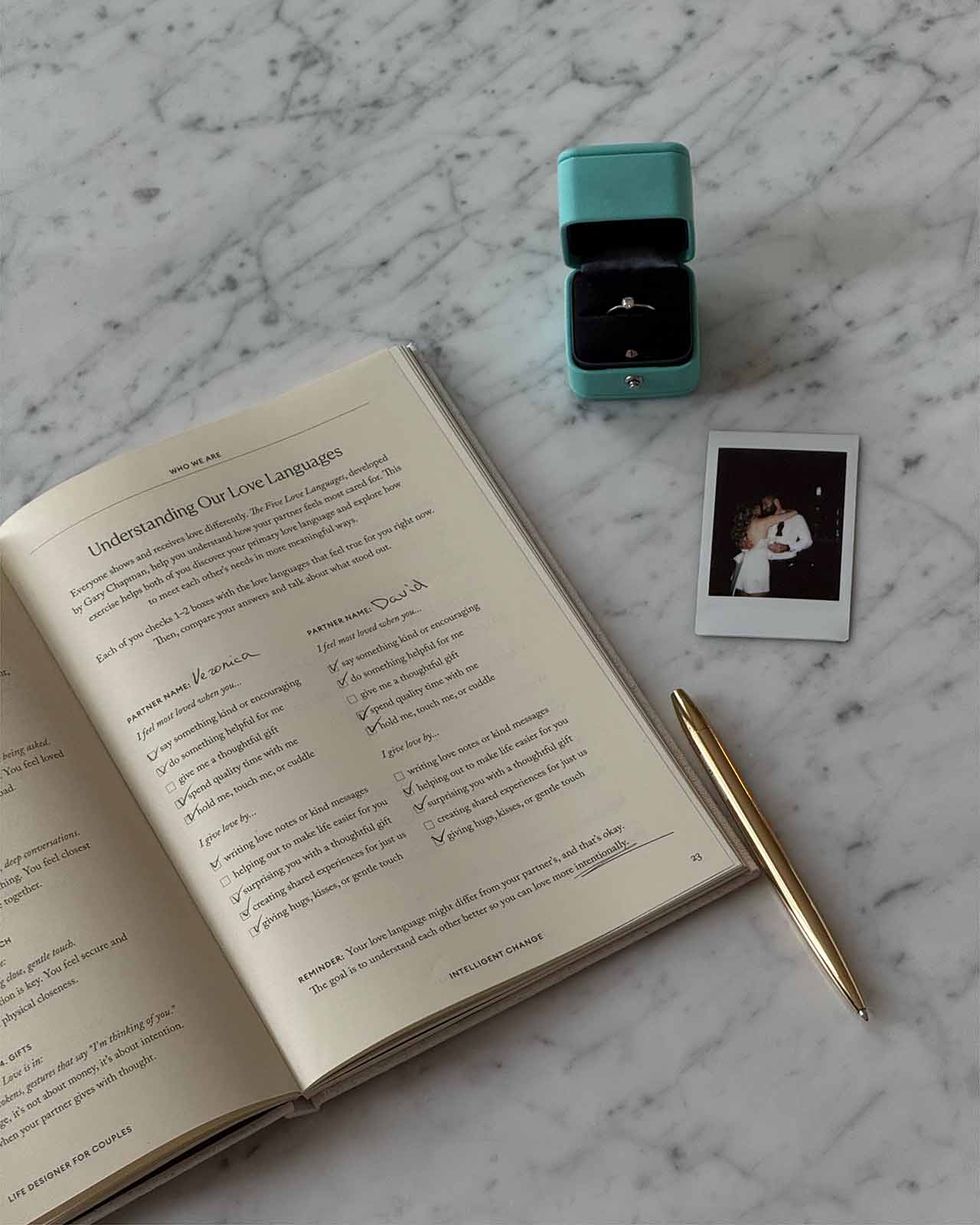 Open book with a pen, teal ring box, and Polaroid photo on a marble surface