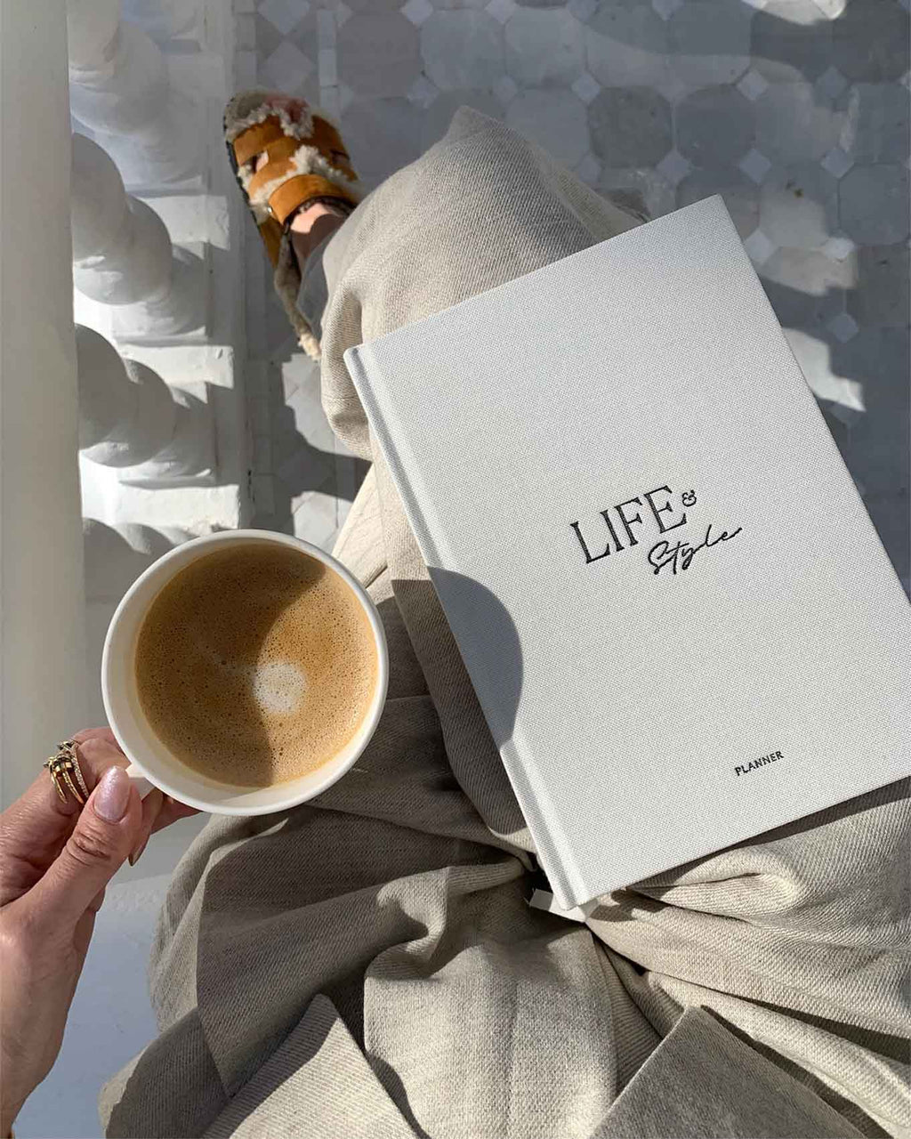 Person holding a cup of coffee with a 'Life & Style' planner on their lap, sitting on a stone bench.
