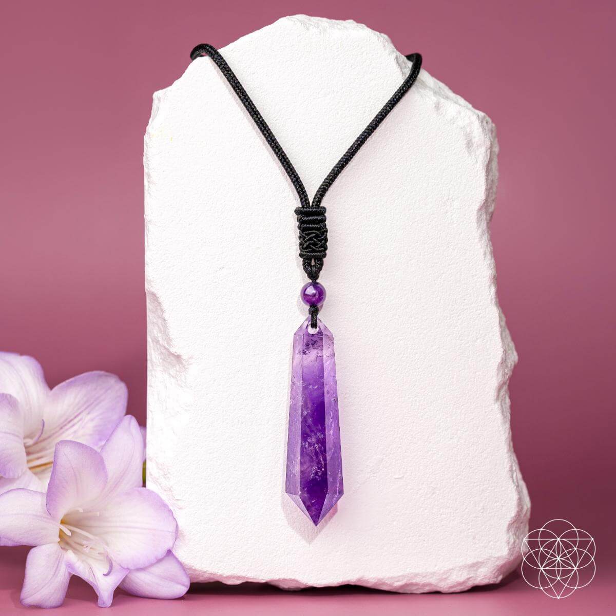 A purple crystal point pendant necklace on a black cord, displayed on a white stone block with purple flowers in the foreground against a pink background.