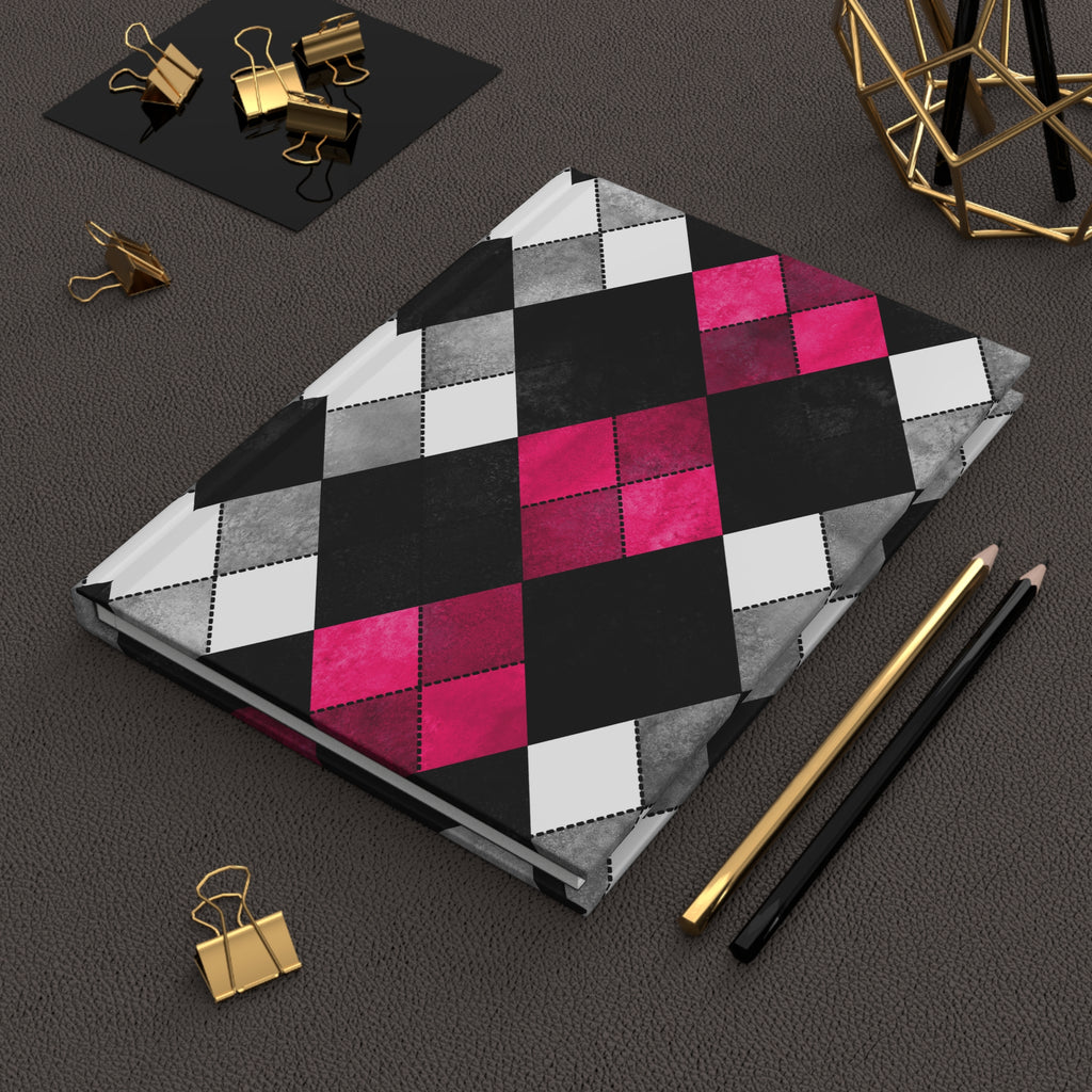 A notebook with a geometric argyle pattern in black, gray, white, and hot pink, styled on a dark textured surface with gold and black office accessories.