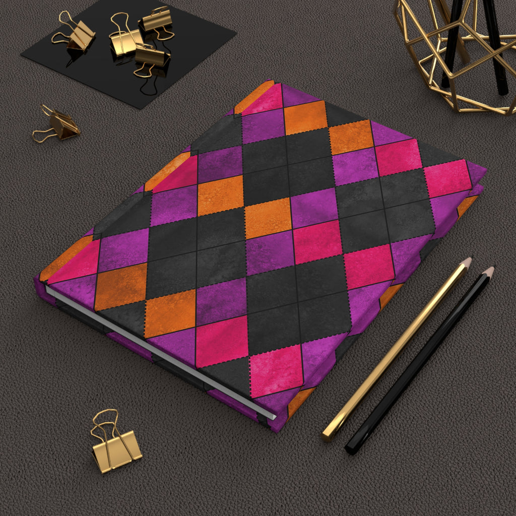 A hardcover notebook with a colorful argyle pattern in purple, orange, and black, styled on a dark textured surface with gold and black office accessories.