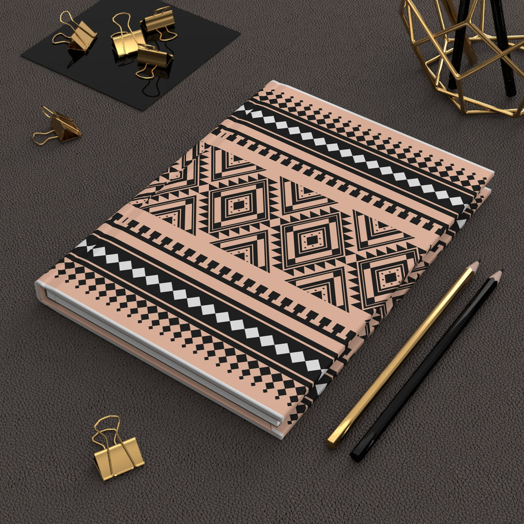 A notebook with a geometric tribal pattern in black, white, and dusty pink, styled on a dark textured surface with gold and black office accessories.