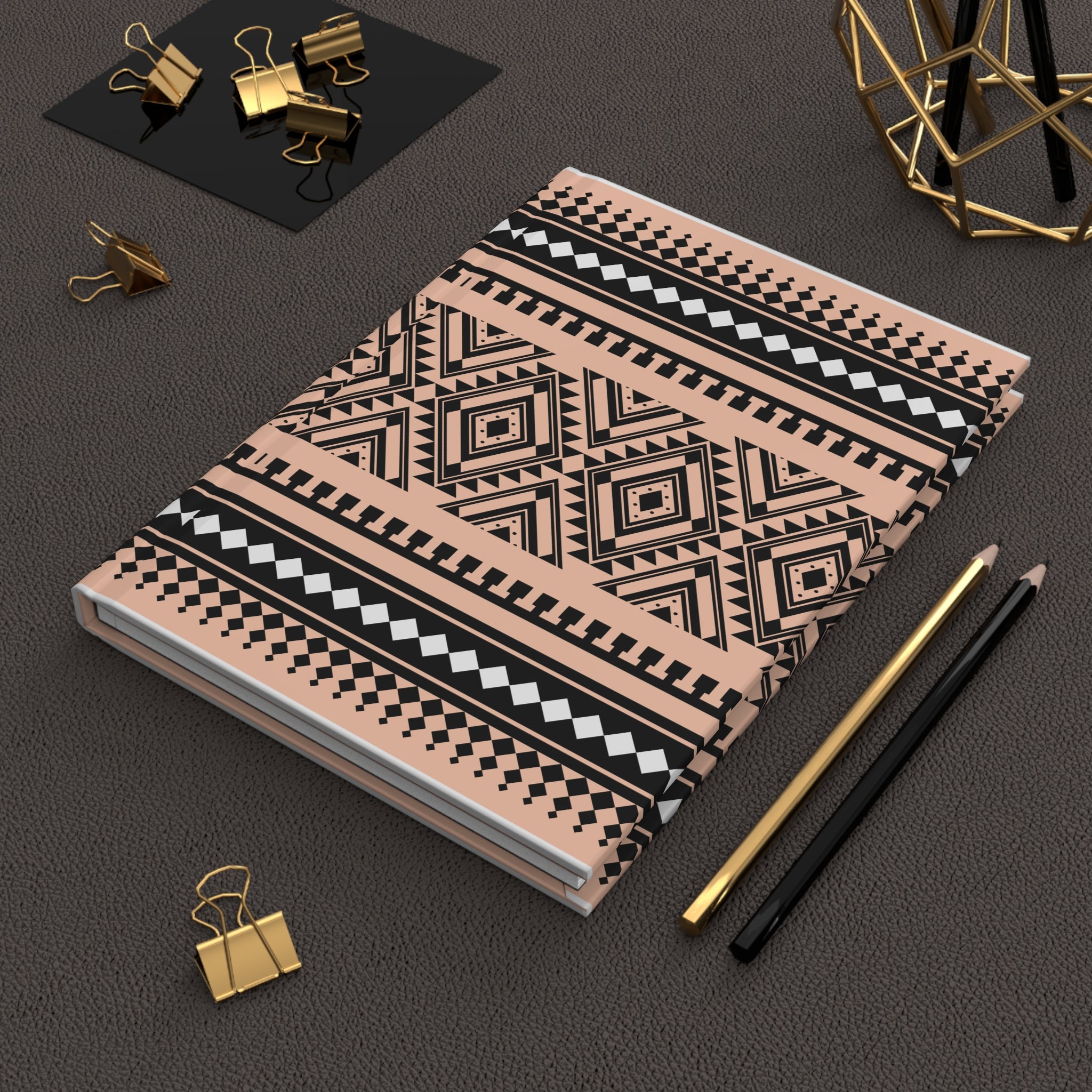 A notebook with a geometric tribal pattern in black, white, and dusty pink, styled on a dark textured surface with gold and black office accessories.
