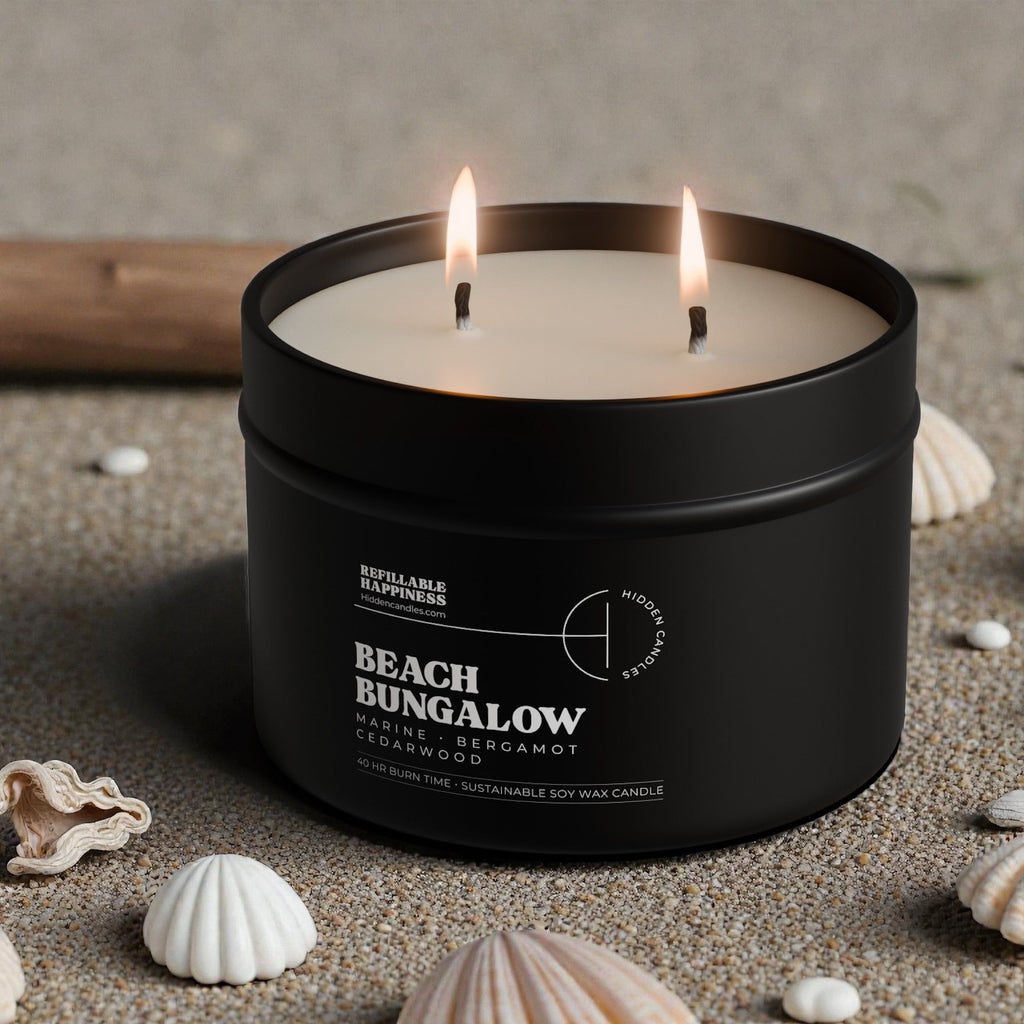 Black candle jar with two lit candles on a sandy surface with seashells.