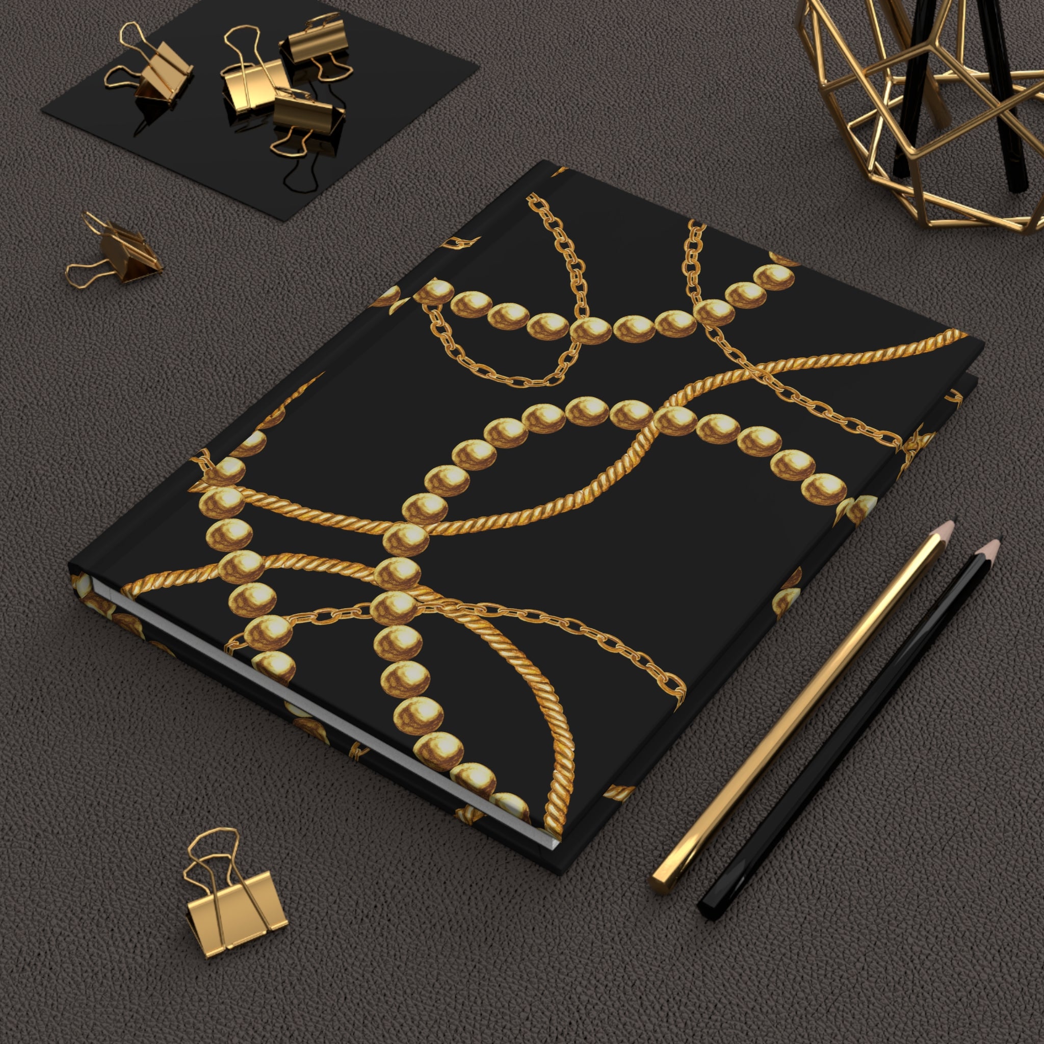 A black notebook or journal with a gold chain and pearl pattern, styled on a dark textured surface with gold binder clips, pencils, and a geometric decor object.