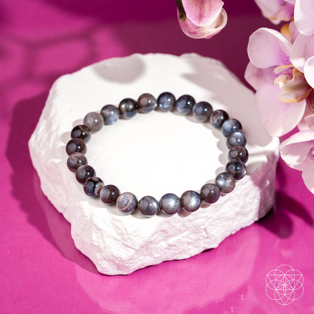 A beaded bracelet made of round gray-blue marbled beads, displayed on a white stone pedestal against a vibrant magenta background with pink orchid flowers in the corner.