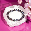 A beaded bracelet made of round gray-blue marbled beads, displayed on a white stone pedestal against a vibrant magenta background with pink orchid flowers in the corner.