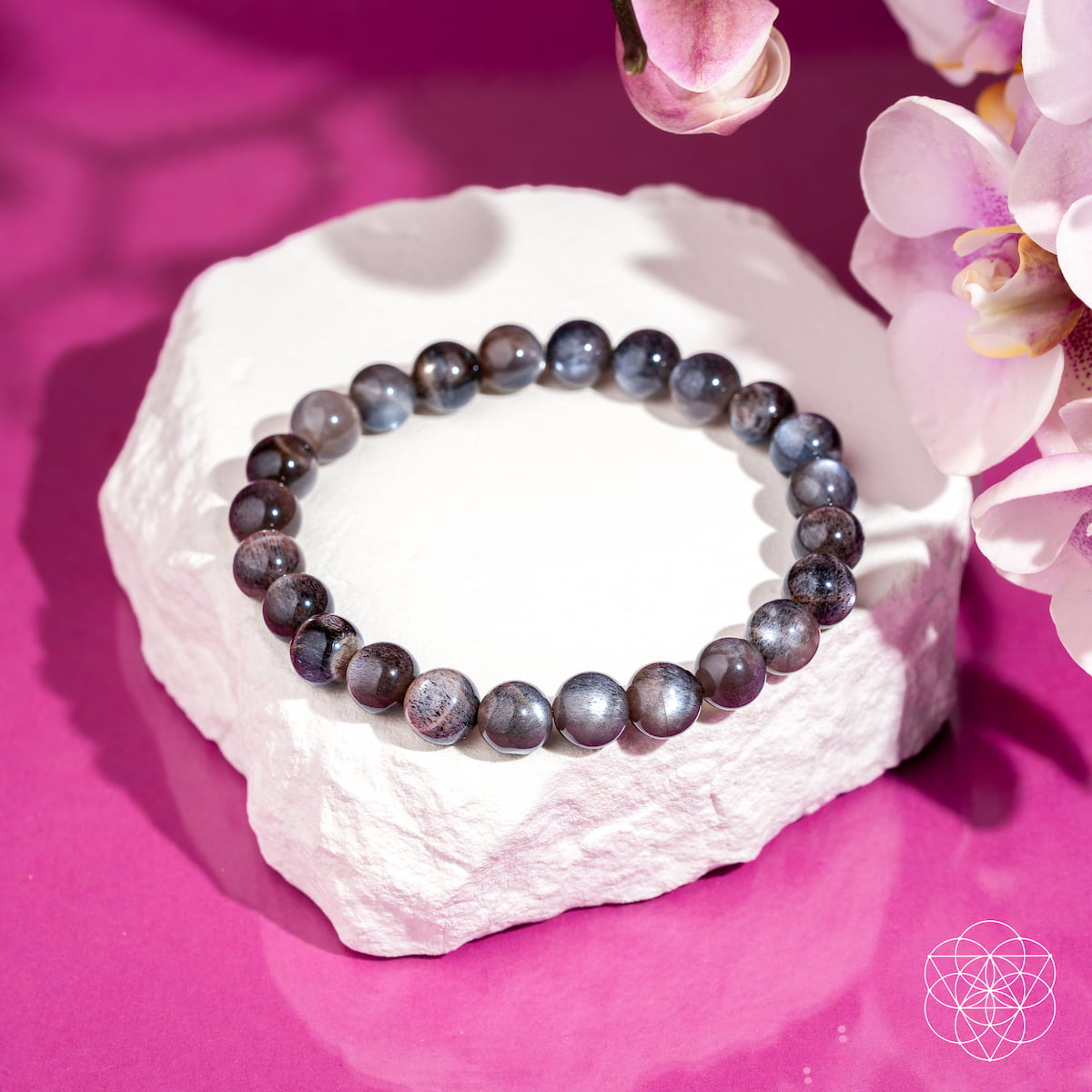 A beaded bracelet made of round gray-blue marbled beads, displayed on a white stone pedestal against a vibrant magenta background with pink orchid flowers in the corner.
