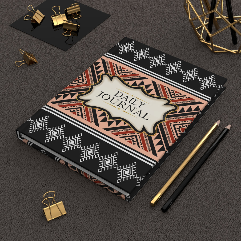A decorative daily journal with a black, beige, and terracotta geometric patterned cover, styled on a dark textured surface with gold and black pencils, binder clips, and a gold geometric decor piece.
