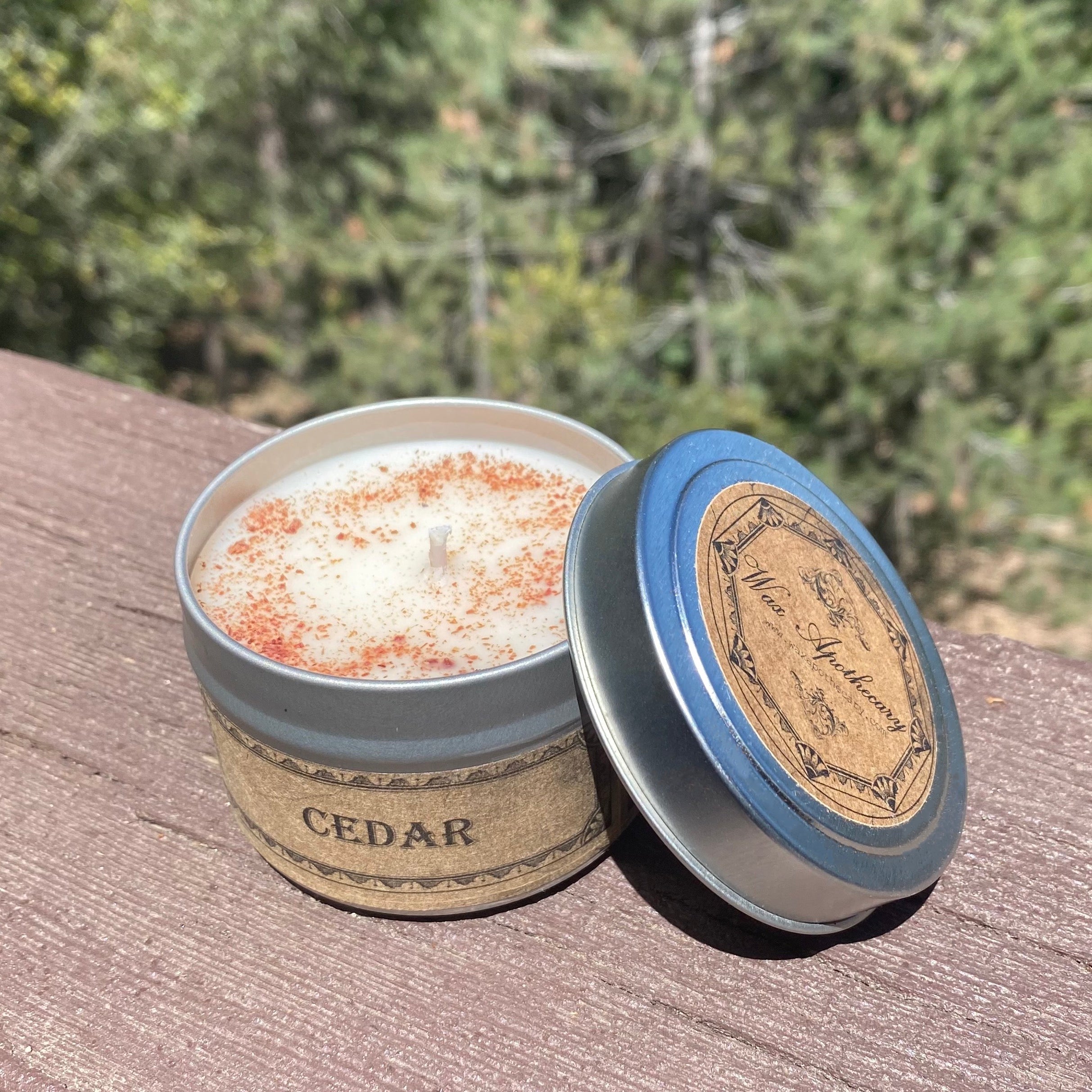 Cedar-scented candle in a tin with a wooden lid on a wooden surface with a forest background.