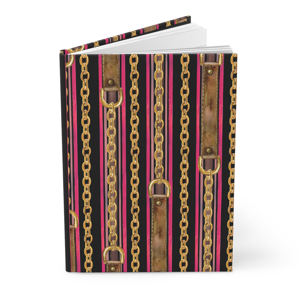 A hardcover notebook or journal standing upright, featuring a bold vertical pattern of gold chains, distressed brown leather straps, and bright pink stripes over a black background.