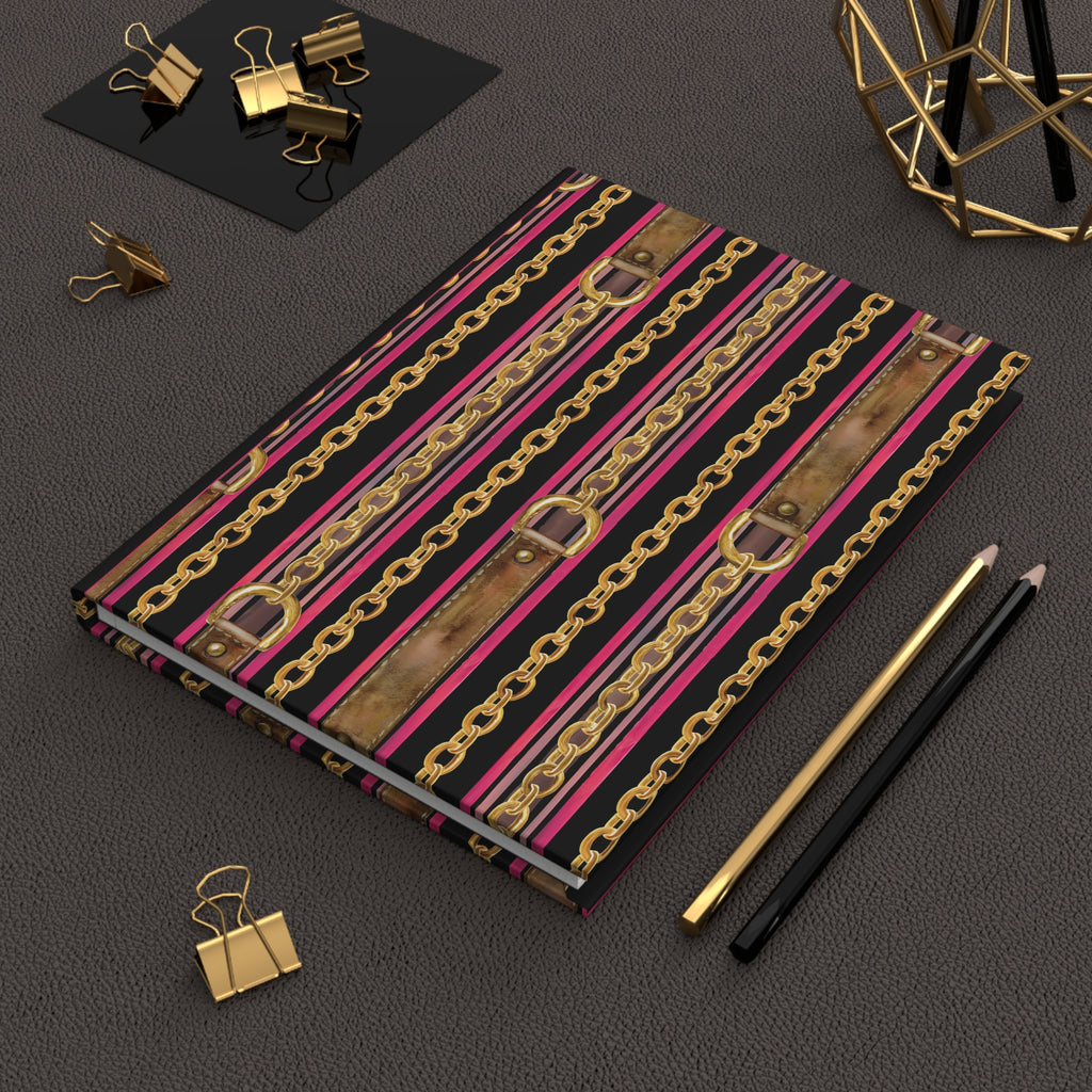 A notebook with a bold pattern of gold chains, pink and brown faux-leather straps, and black stripes, styled on a dark textured surface with gold and black office accessories.