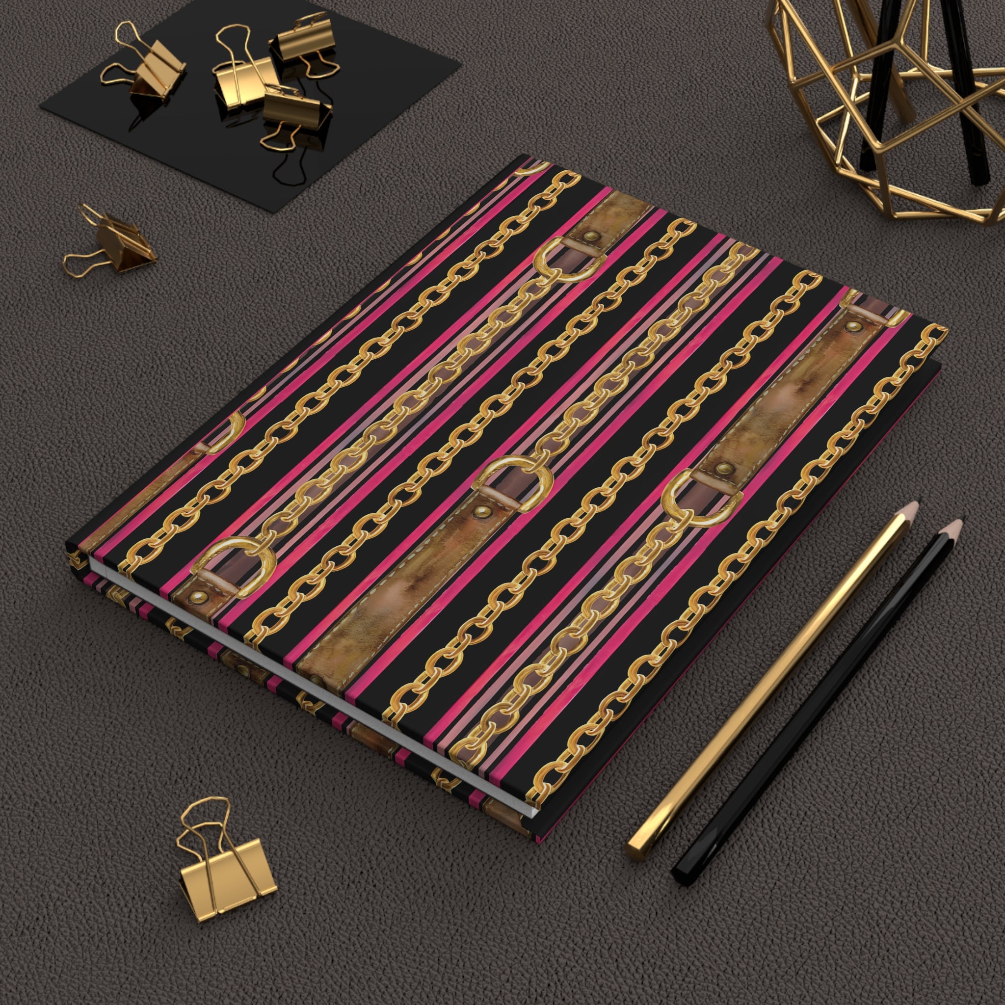 A notebook with a bold pattern of gold chains, pink and brown faux-leather straps, and black stripes, styled on a dark textured surface with gold and black office accessories.