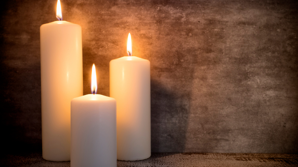 Three lit white pillar candles of varying heights arranged on a textured surface against a dark, rustic brown background.