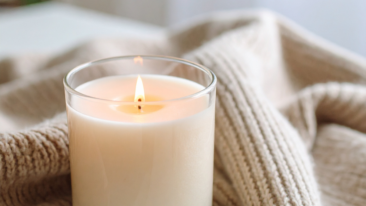 A lit white candle in a clear glass jar, resting on a cozy beige knit blanket.