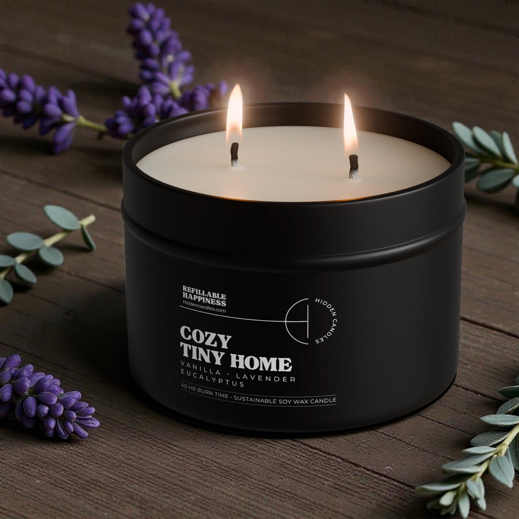 Black candle jar with two lit candles on a wooden surface with lavender and green leaves.