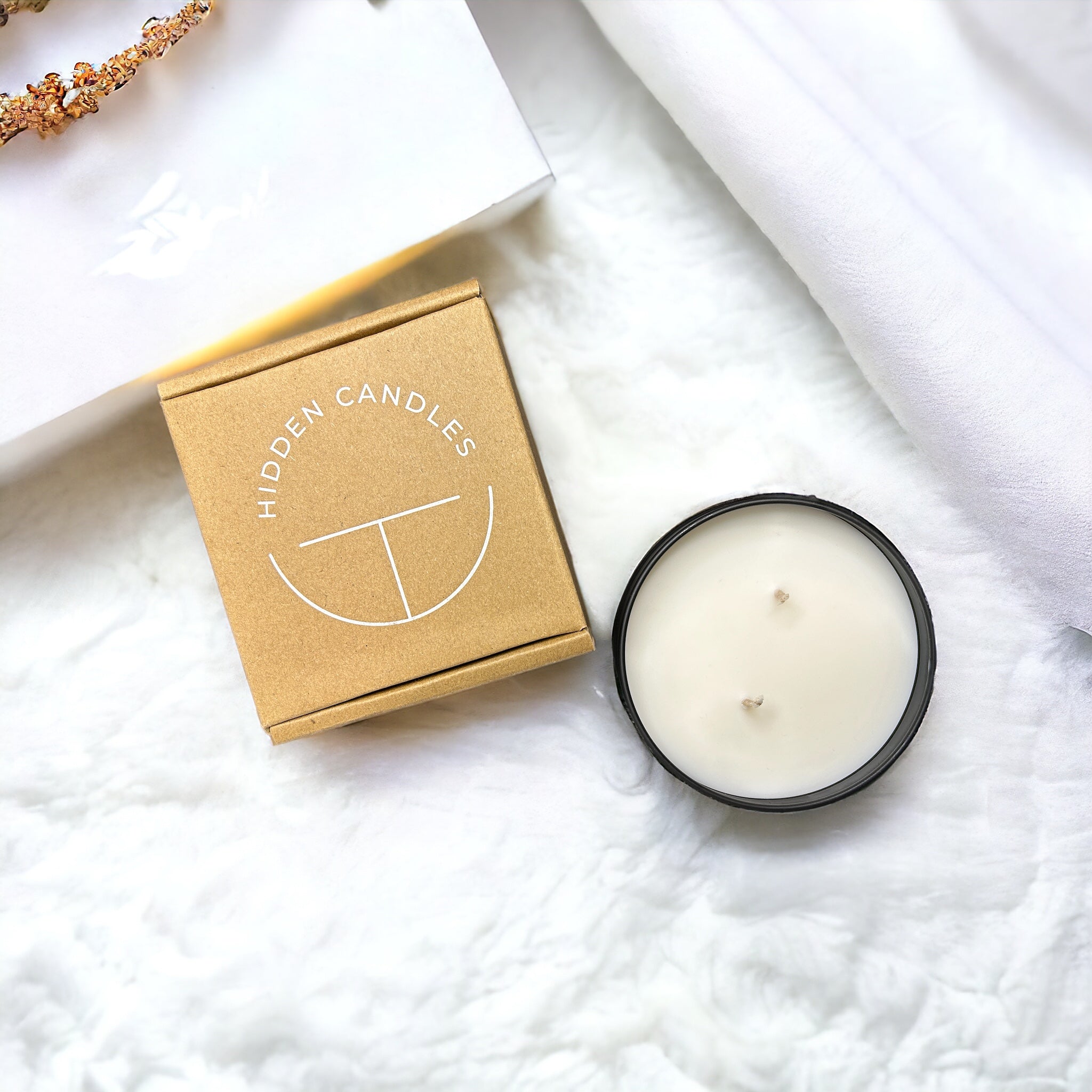 White candle in a black container with a 'Hidden Candles' box on a white surface