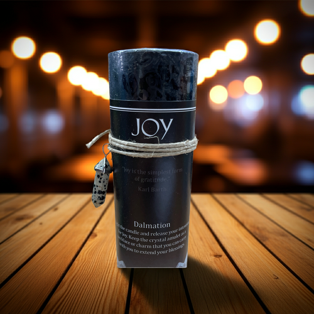 A rolled black crystal candle wrapped in twine with a small hanging tag, standing on a wooden surface against a blurred warm bokeh background. The label reads JOY and Dalmation.