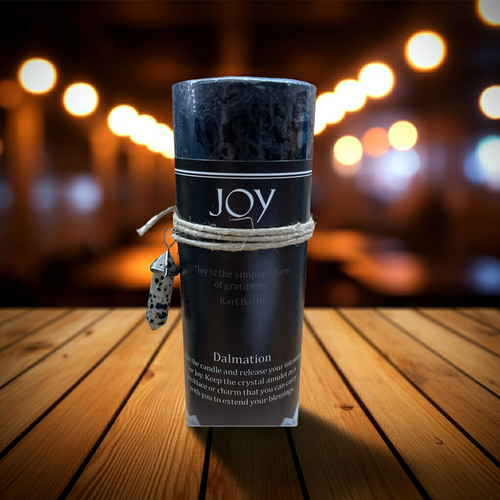 A rolled black crystal candle wrapped in twine with a small hanging tag, standing on a wooden surface against a blurred warm bokeh background. The label reads JOY and Dalmation.