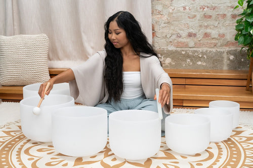 Woman playing white tibetan singing bowls in a room with a brick wall and plant.
