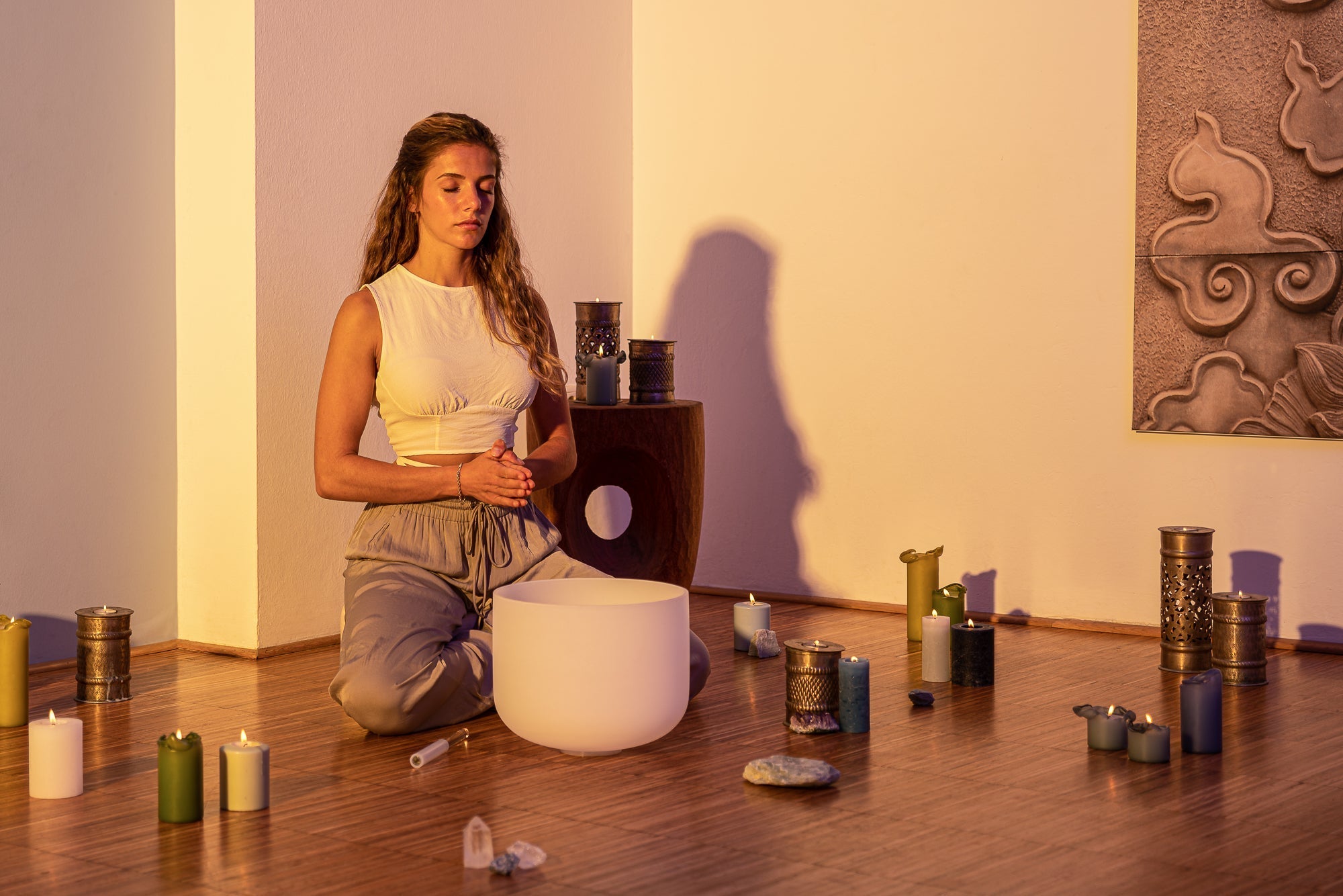 Woman kneeling in front of a white crystal bowl in a room with candles and crystals.