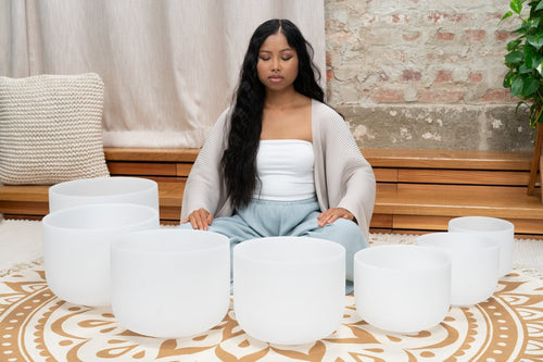 Woman meditating in front of white crystal bowls on a patterned rug.
