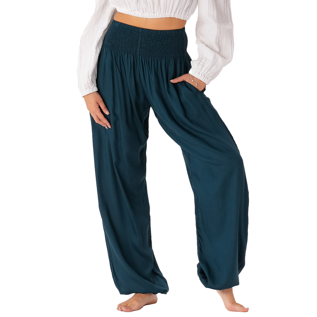 A person modeling loose, wide-leg harem-style pants in a deep teal-blue color, paired with a white long-sleeve crop top. The pants feature a gathered elastic waist, side pockets, and a relaxed drape, shown against a plain white background.