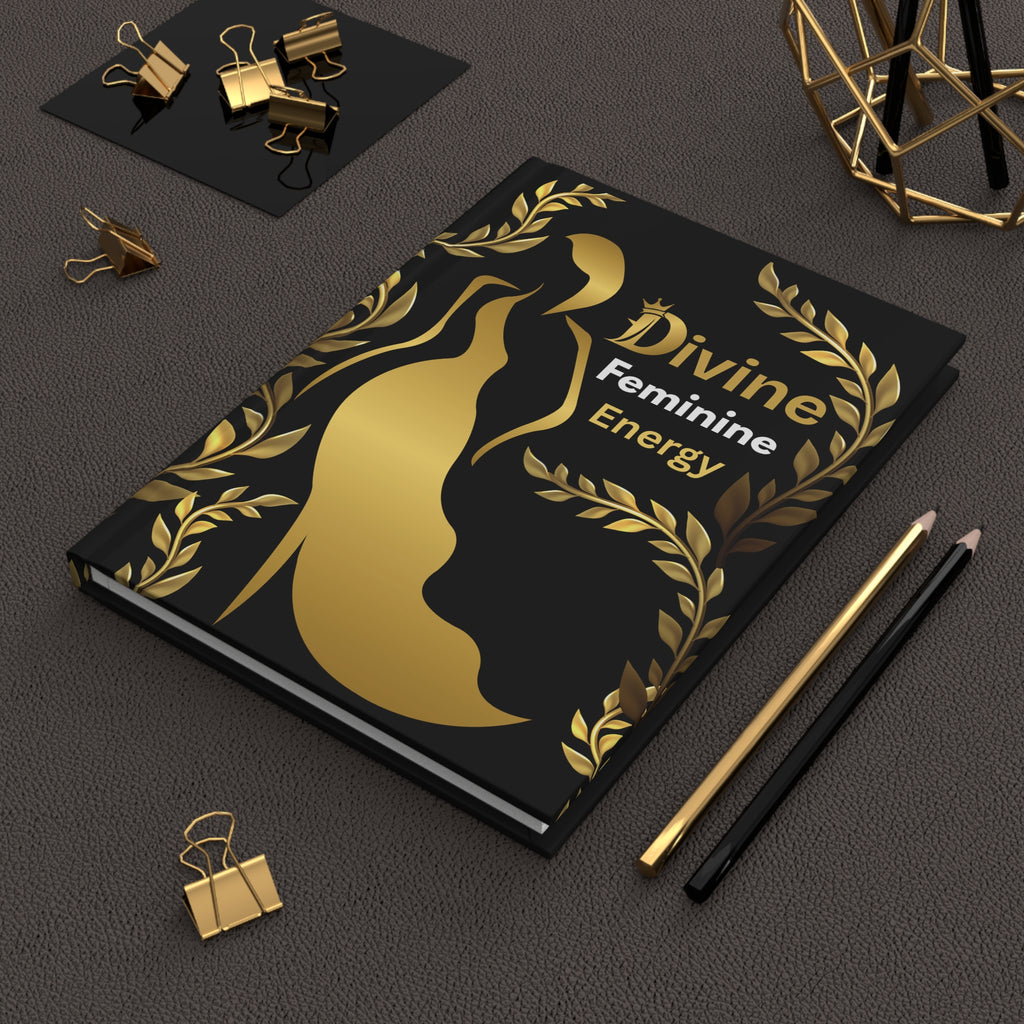 A black notebook with a gold metallic silhouette of a female figure and laurel wreath, featuring the text 'Divine Feminine Energy'. The notebook is styled on a dark textured surface with gold and black pencils, binder clips, and a geometric gold object in the background.
