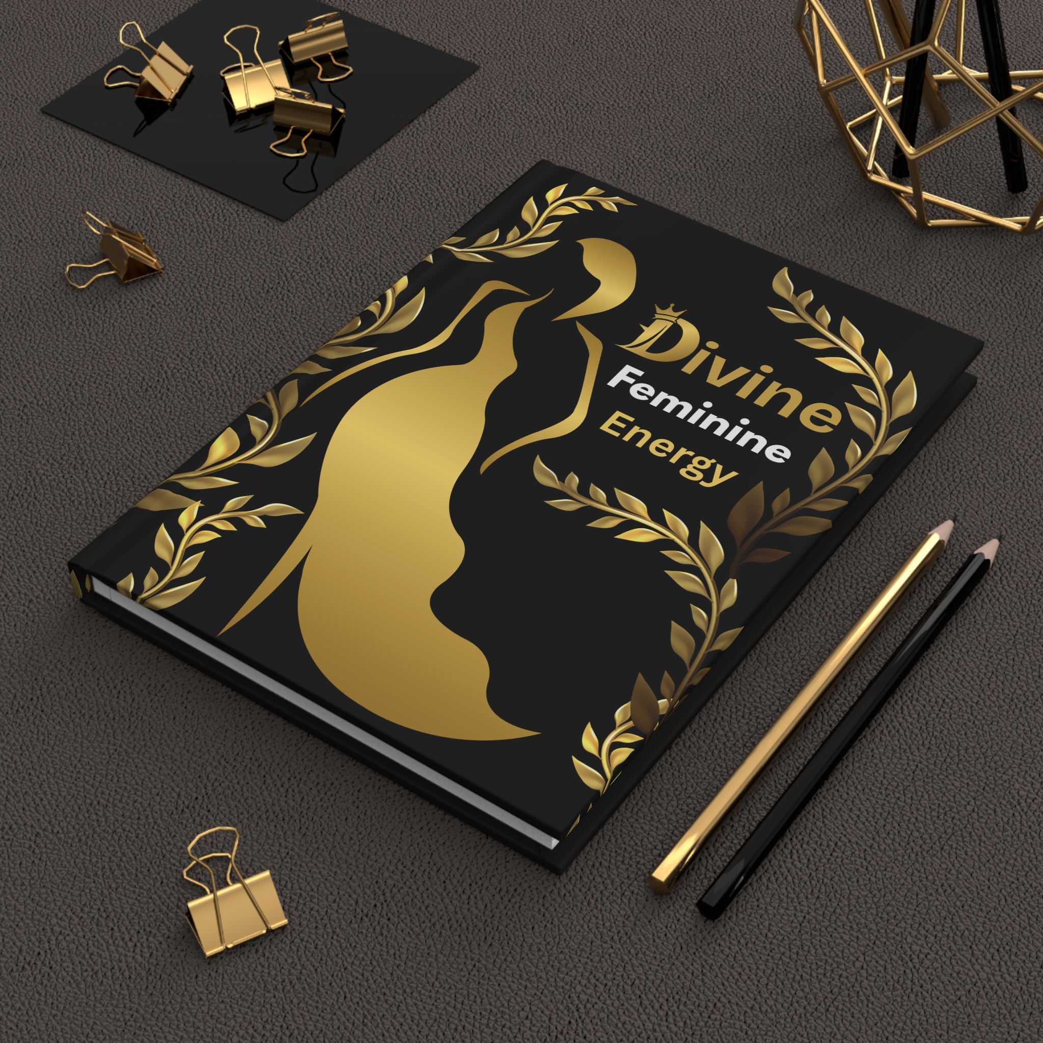 A black notebook with a gold metallic silhouette of a female figure and laurel wreath, featuring the text 'Divine Feminine Energy'. The notebook is styled on a dark textured surface with gold and black pencils, binder clips, and a geometric gold object in the background.