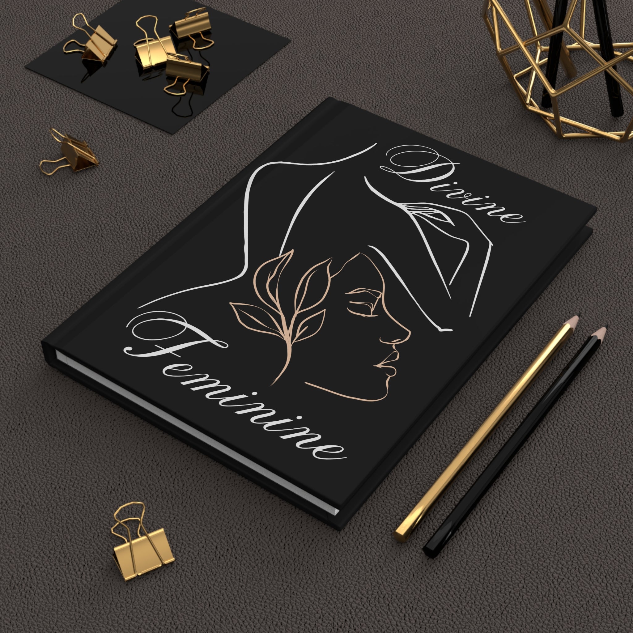 A black hardcover notebook with elegant white and gold line art of a woman's profile and the words 'Divine Feminine' on the cover, styled on a dark textured surface with gold and black office accessories.