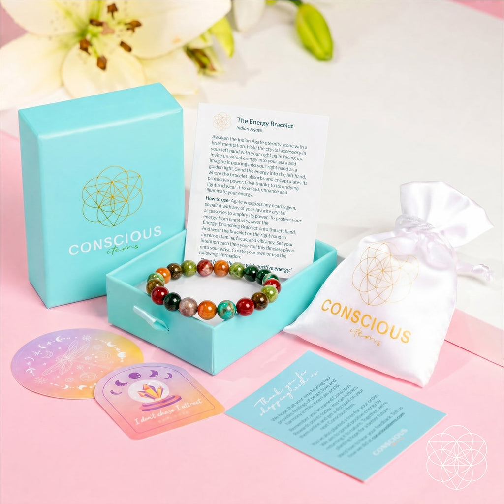 A Conscious Energy brand energy bracelet set displayed on a pink background. The set includes a colorful beaded bracelet, a light blue presentation box, a white drawstring pouch, and printed cards and coasters.