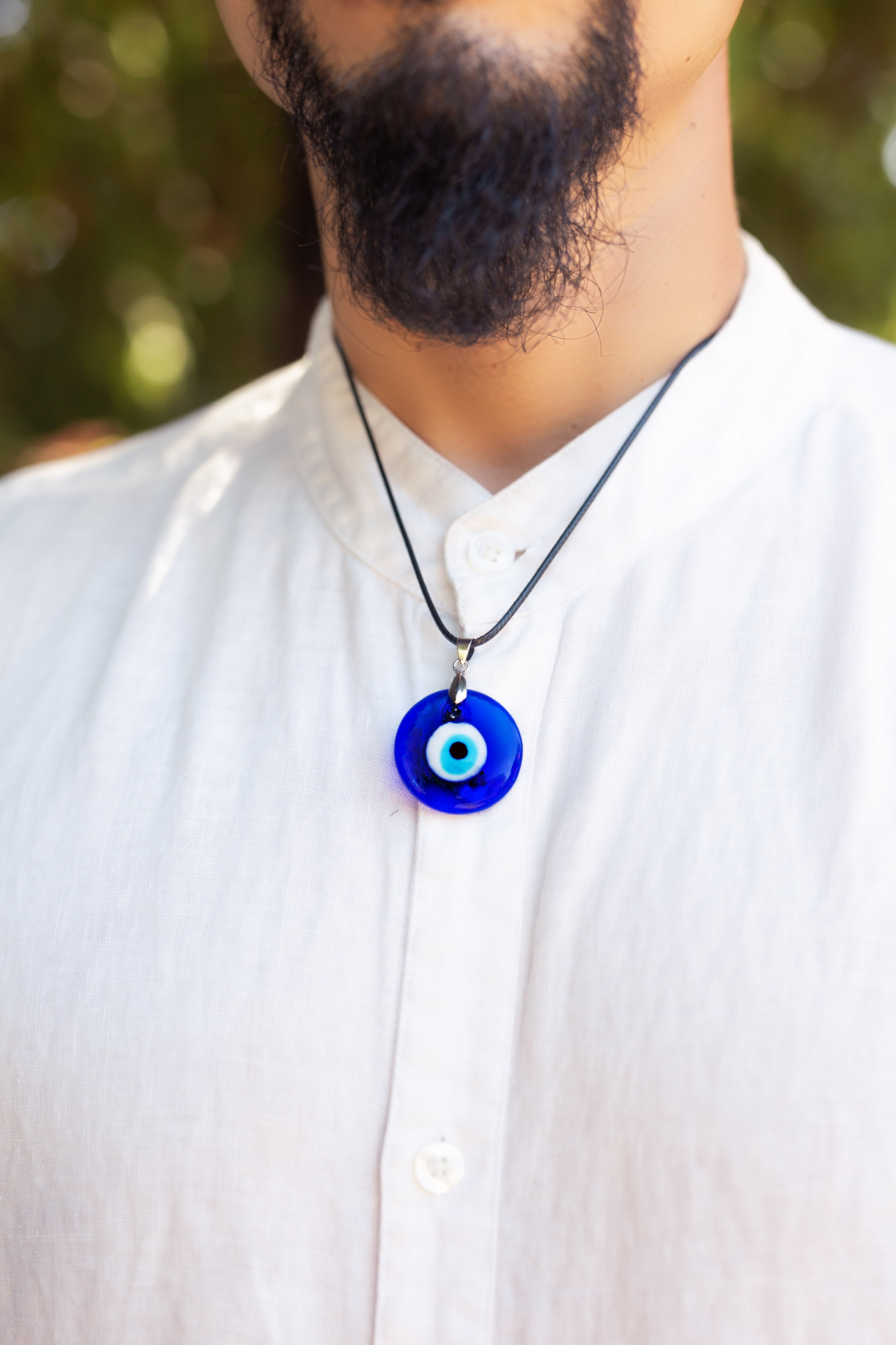 A close-up of a man wearing a white linen shirt and a black cord necklace with a blue glass evil eye pendant.