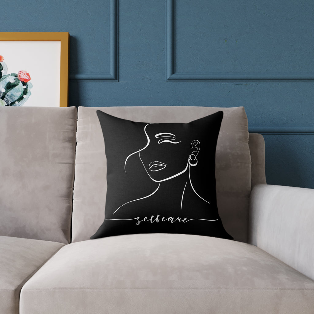 Black pillow with a woman's face and 'selfcare' text on a gray couch against a blue wall.