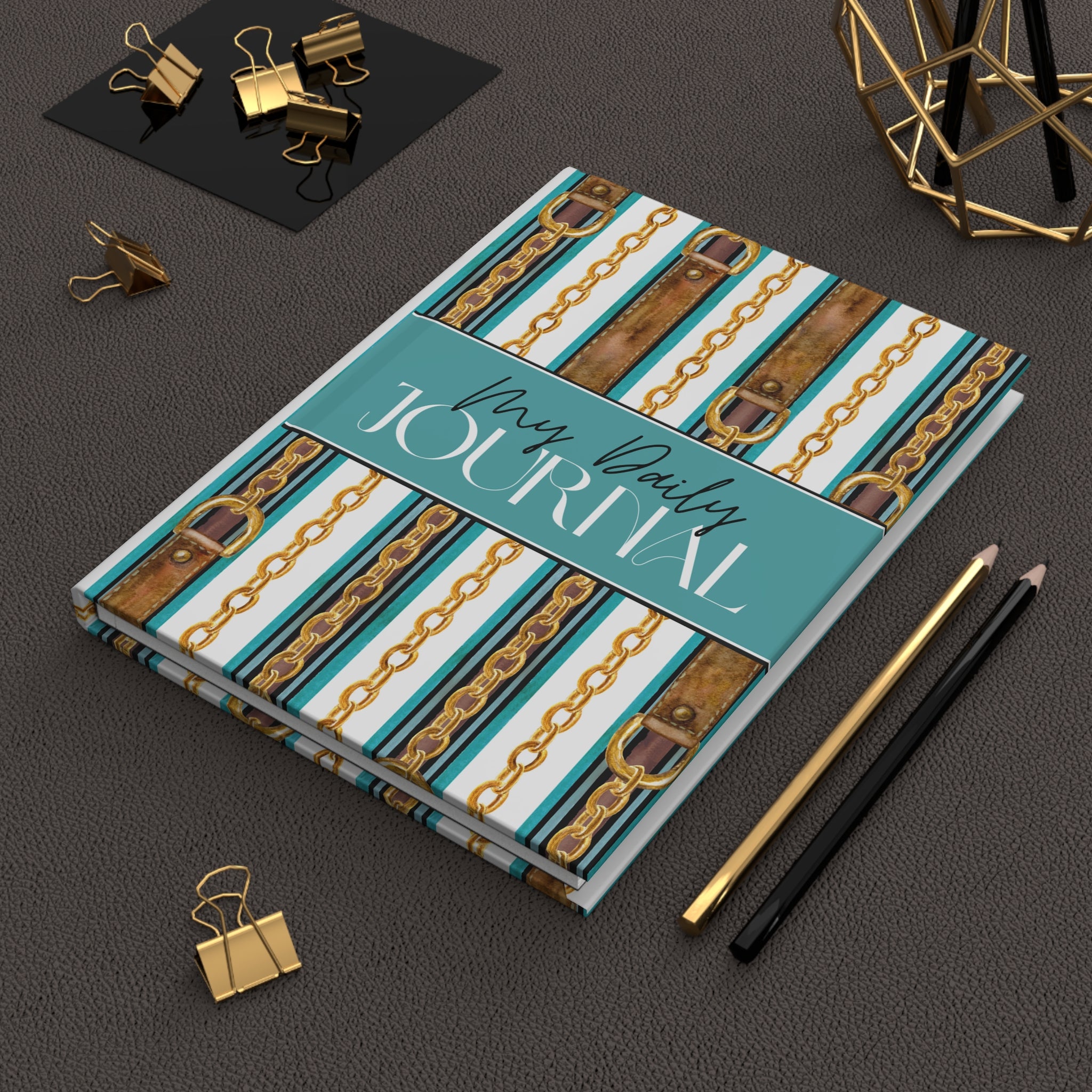 A hardcover daily journal with a teal, white, and brown chain-and-strap patterned cover, styled on a dark textured surface with gold binder clips, two pencils, and a geometric gold decor object.