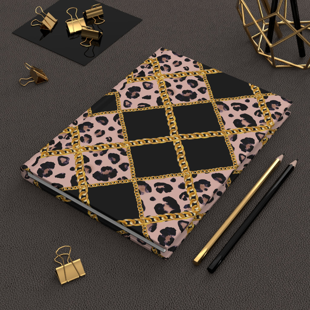 A notebook with a pink leopard print and black diamond pattern separated by gold chain motifs, styled on a dark textured surface with gold and black pencils, binder clips, and a geometric gold decor object.