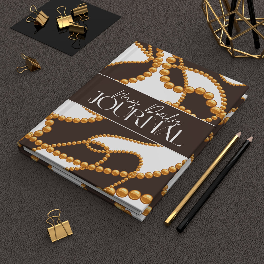 A hardcover journal with a brown and white pearl-patterned cover, styled on a dark textured surface with gold binder clips, pencils, and a geometric decor object.