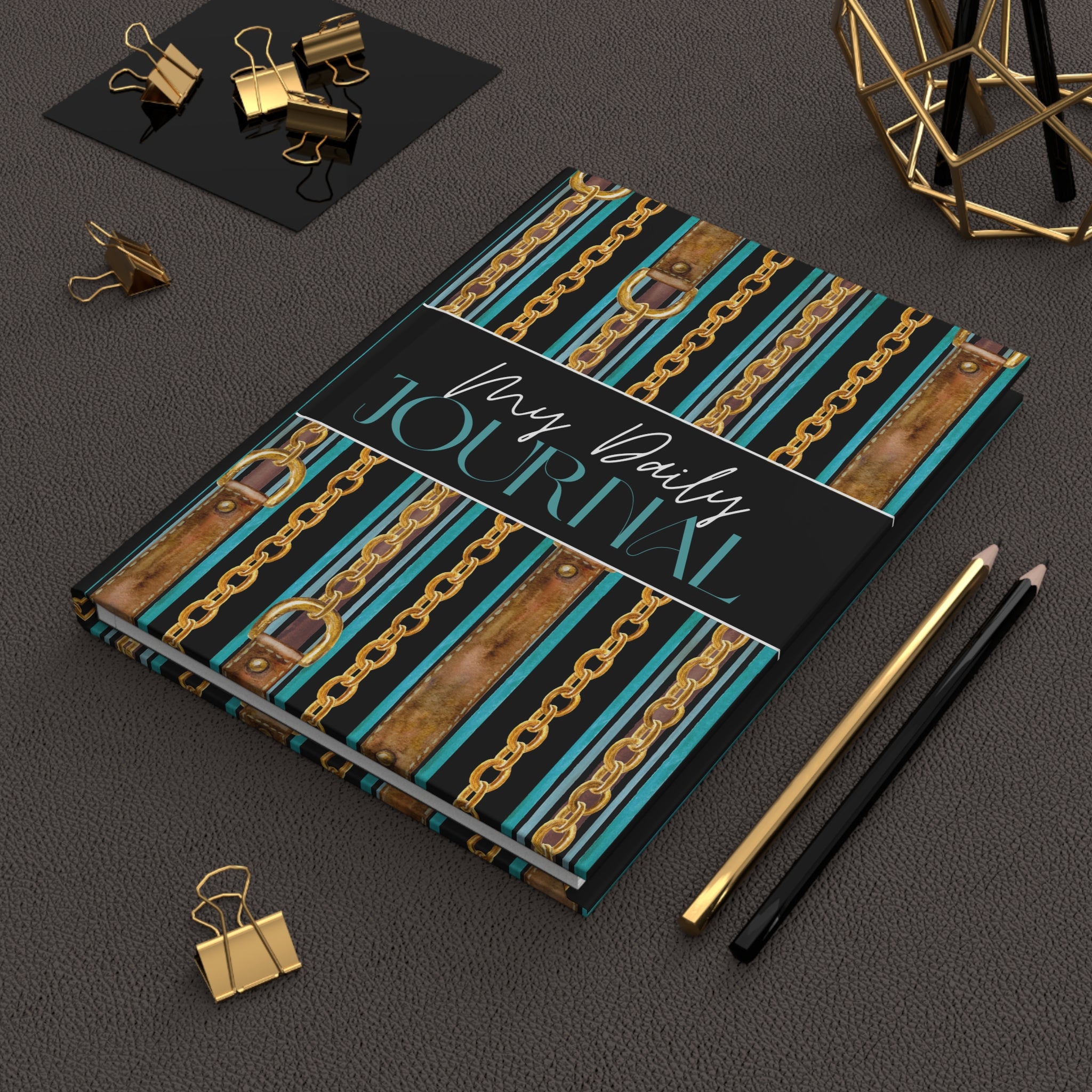 A hardcover journal with a black cover featuring turquoise and brown striped chains, shown in a styled flat-lay with gold binder clips, pencils, and a geometric decor object on a dark textured surface.