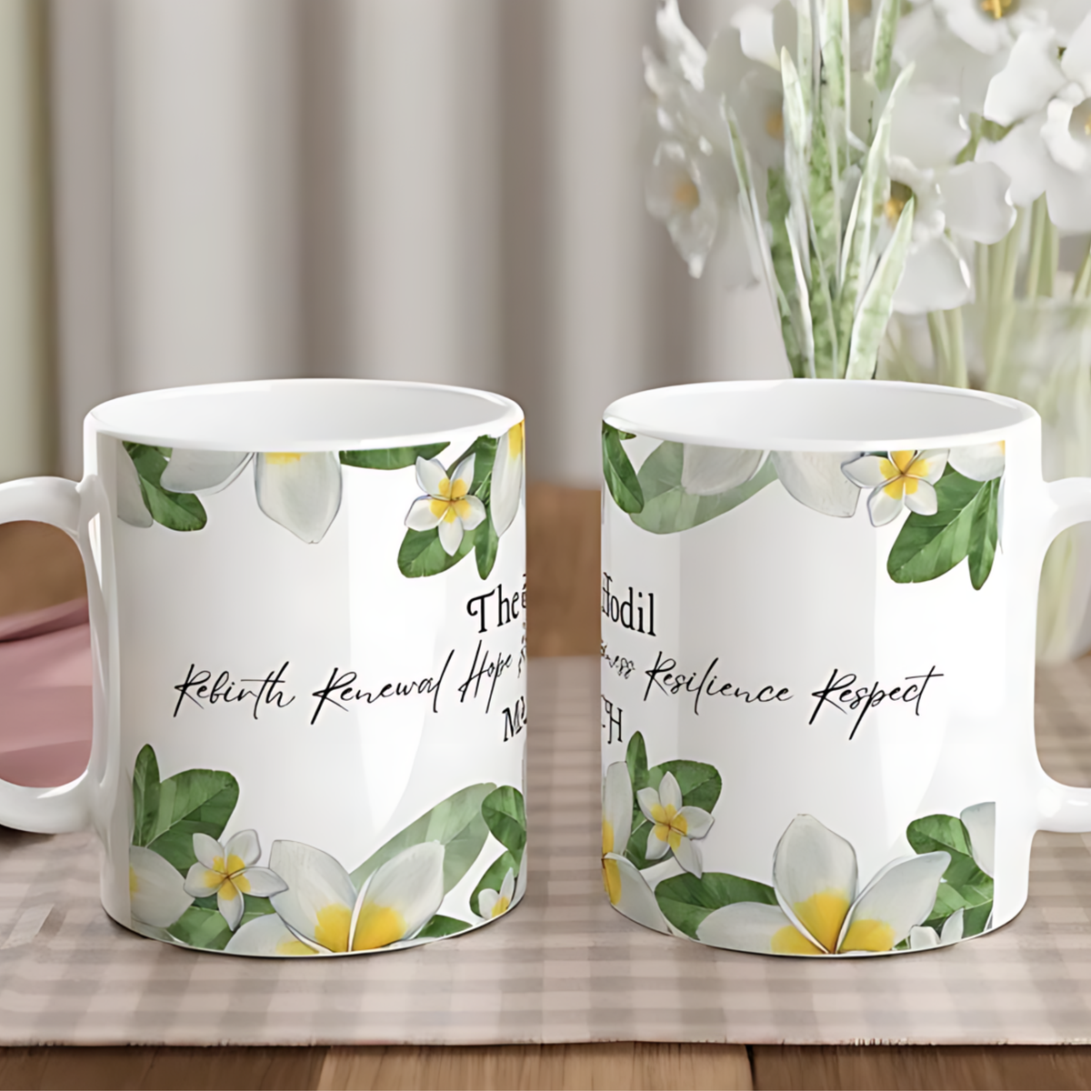 Two white ceramic mugs with a floral print of white plumeria flowers and green leaves, featuring black cursive text, displayed on a checkered tablecloth with a blurred vase of flowers in the background.