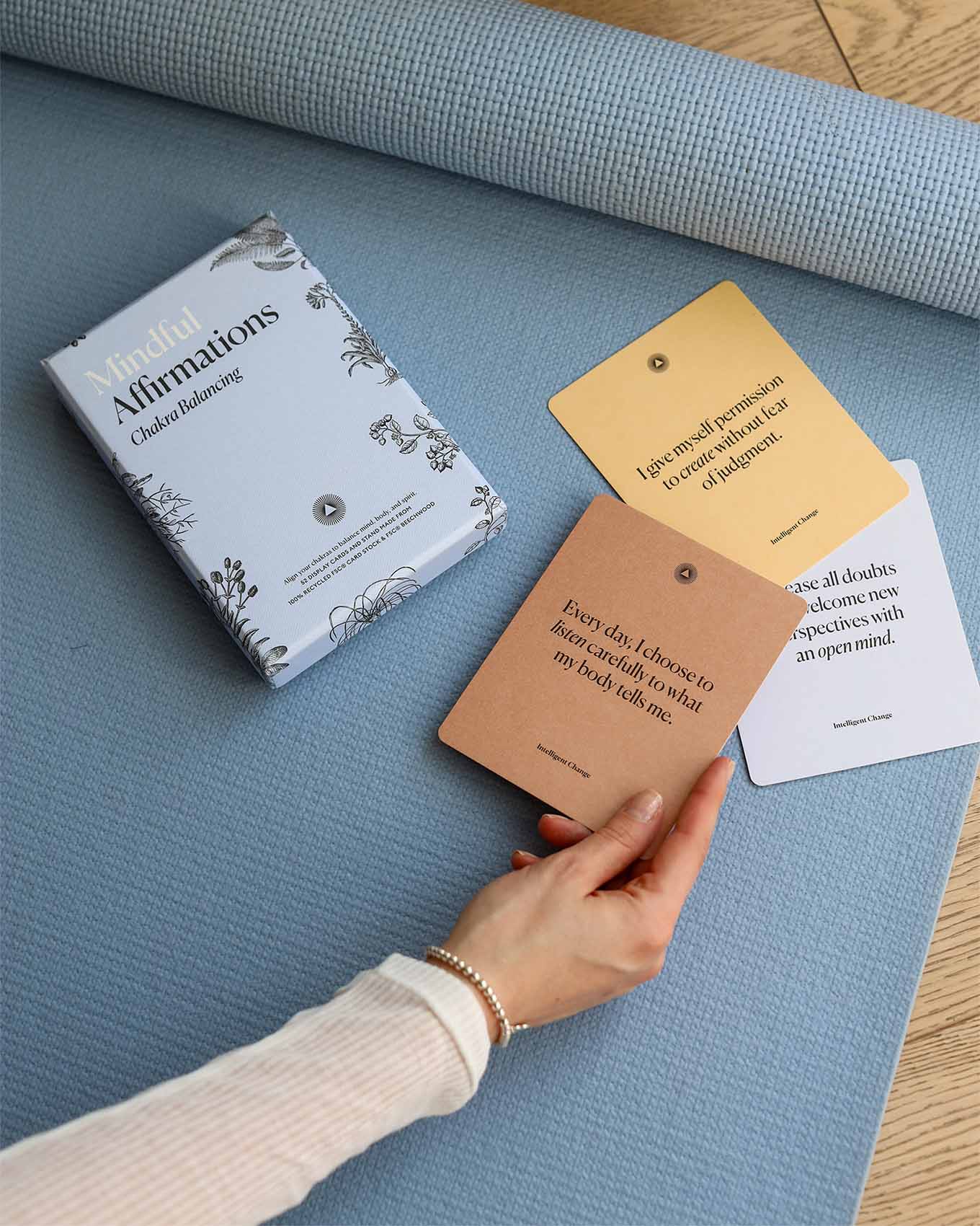 Book titled 'Mindful Affirmations' on a blue mat with a hand holding cards.