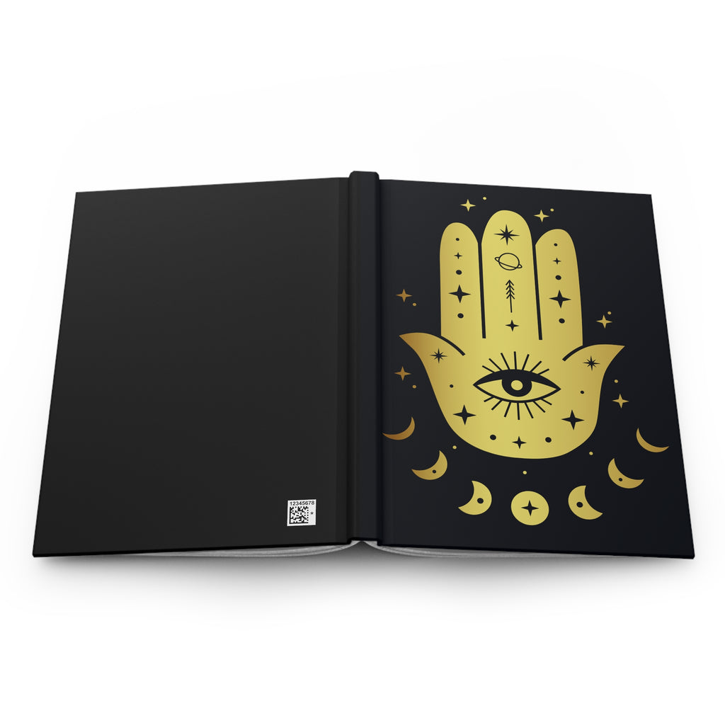 A black hardcover notebook or journal shown open, featuring a large gold Hamsa hand illustration with an eye of providence, stars, and crescent moons on the right page.