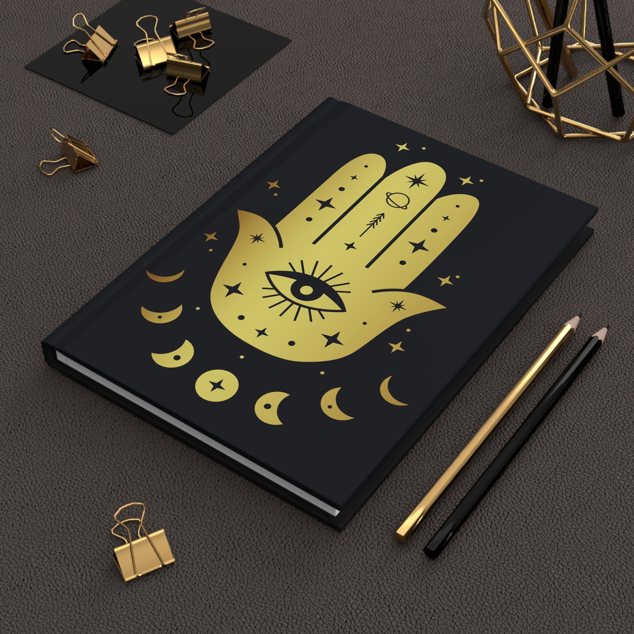 A black hardcover notebook with a gold Hamsa hand design featuring an eye of providence and crescent moons, styled on a dark textured surface with gold and black pencils and binder clips.
