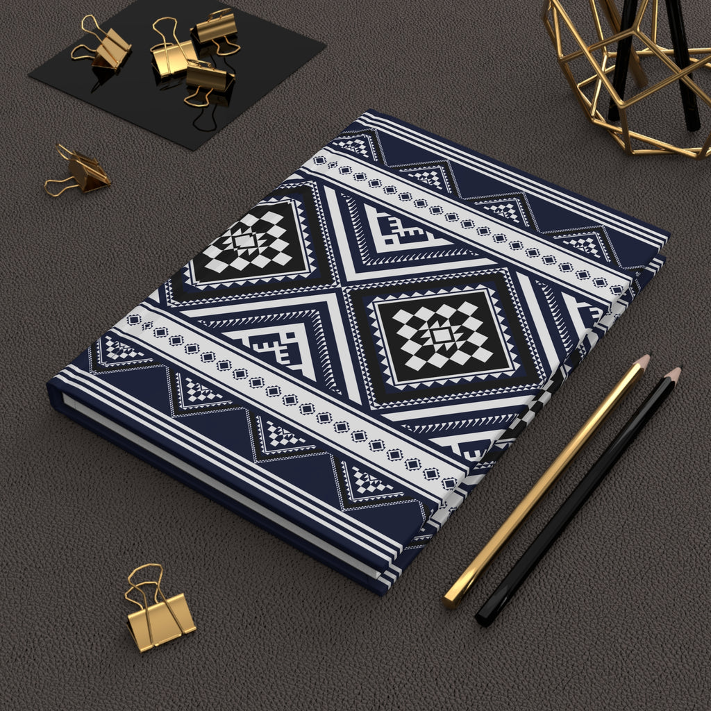 A notebook with a navy blue, white, and black geometric tribal pattern, styled on a dark textured surface with gold binder clips, pencils, and a gold geometric decor piece.