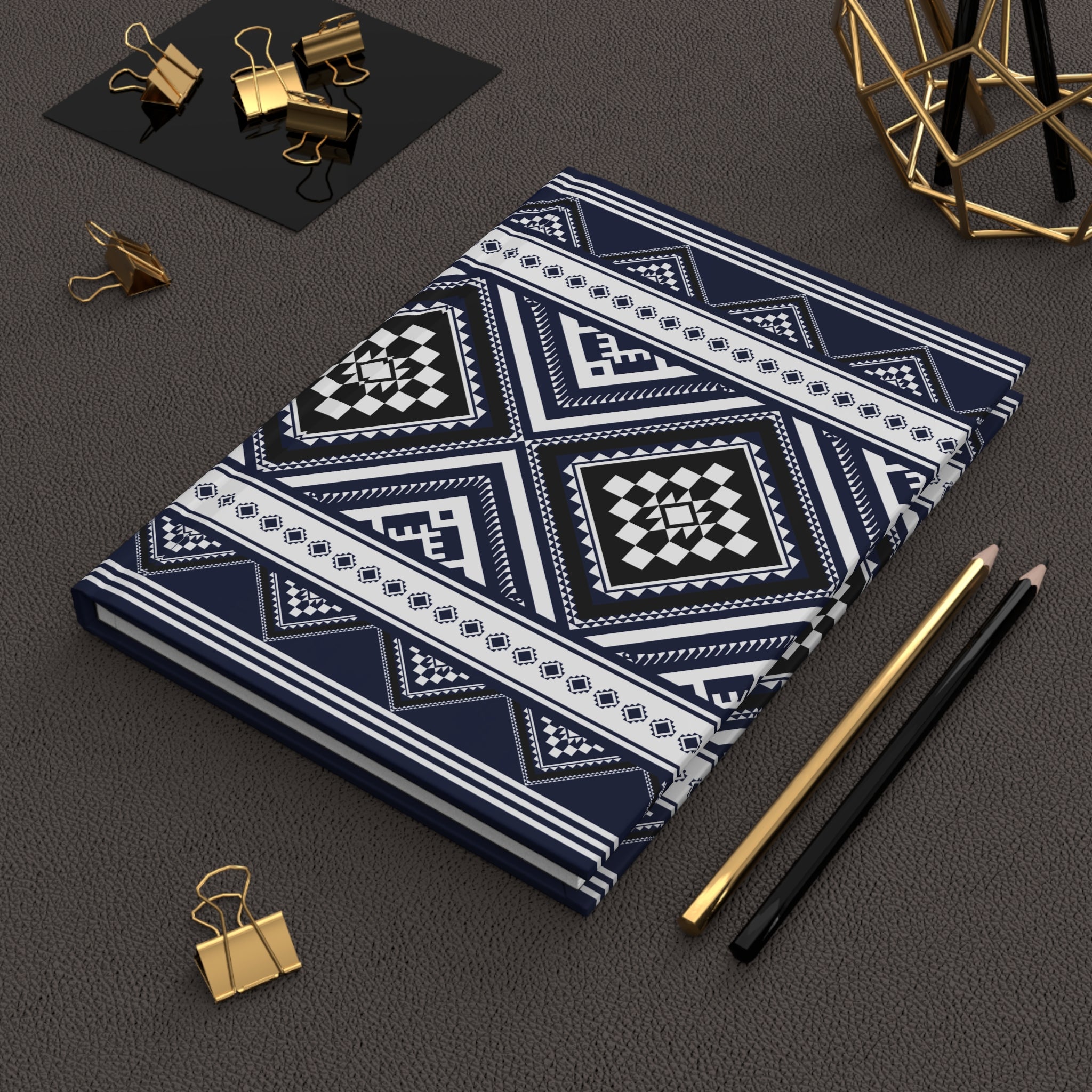 A notebook with a navy blue, white, and black geometric tribal pattern, styled on a dark textured surface with gold binder clips, pencils, and a gold geometric decor piece.