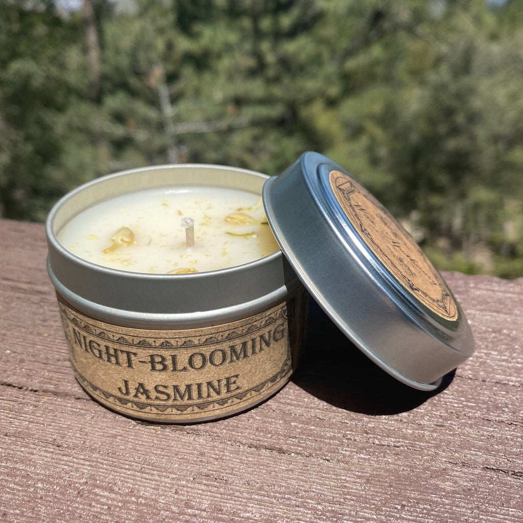 Candle in a tin labeled 'Night-Blooming Jasmine' on a wooden surface with a blurred natural background