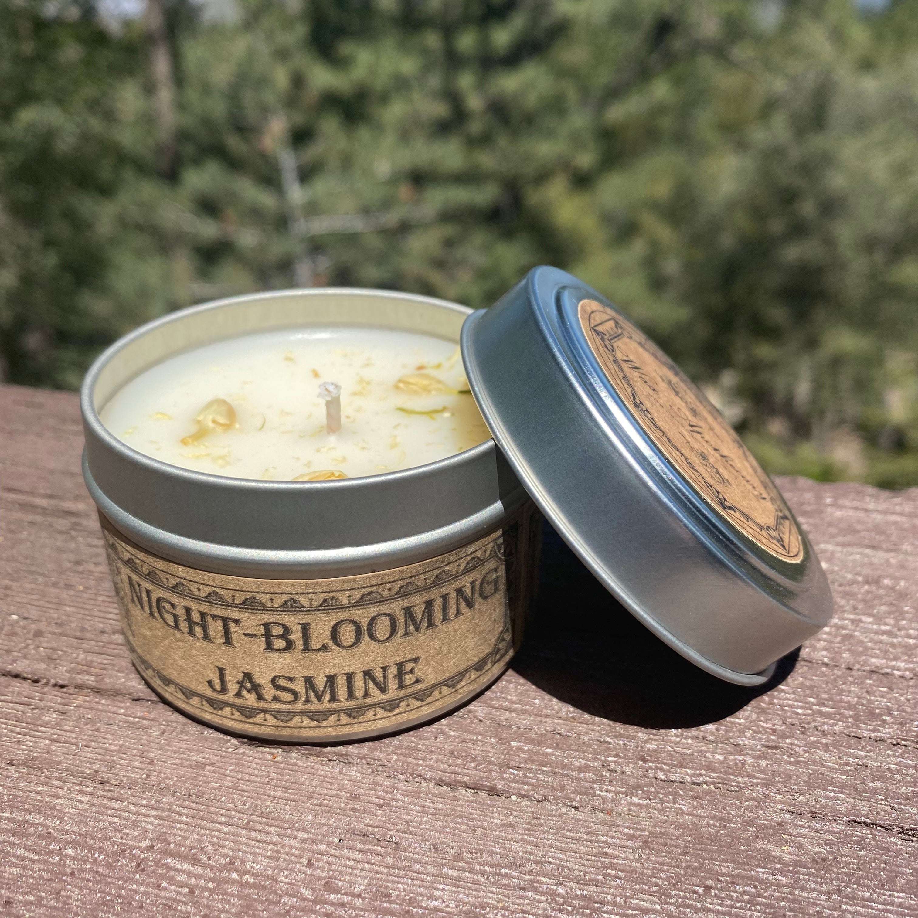 Candle in a tin labeled 'Night-Blooming Jasmine' on a wooden surface with a blurred natural background