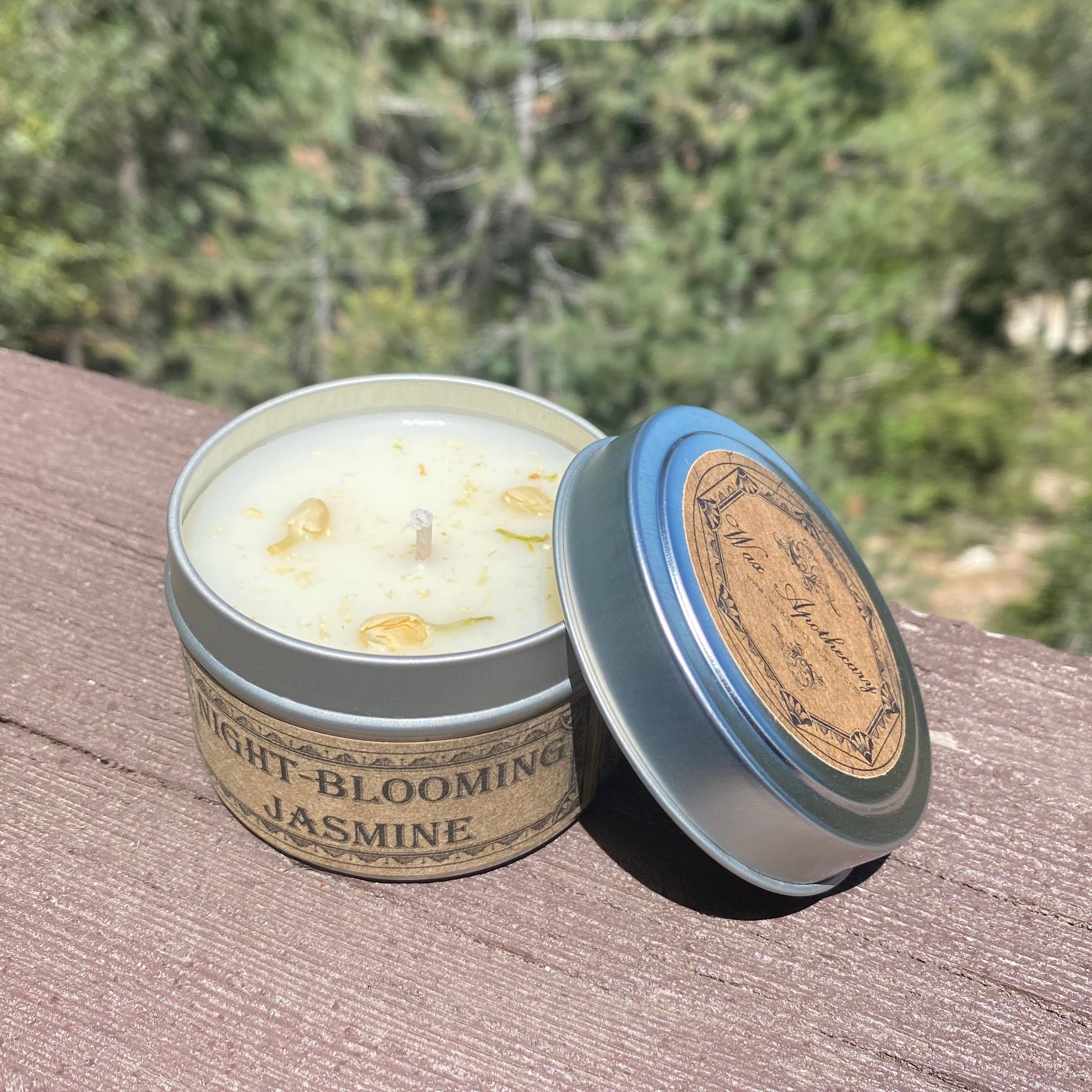 Candle in a tin labeled 'Night Blooming Jasmine' with a wooden lid on a wooden surface with a blurred natural background.
