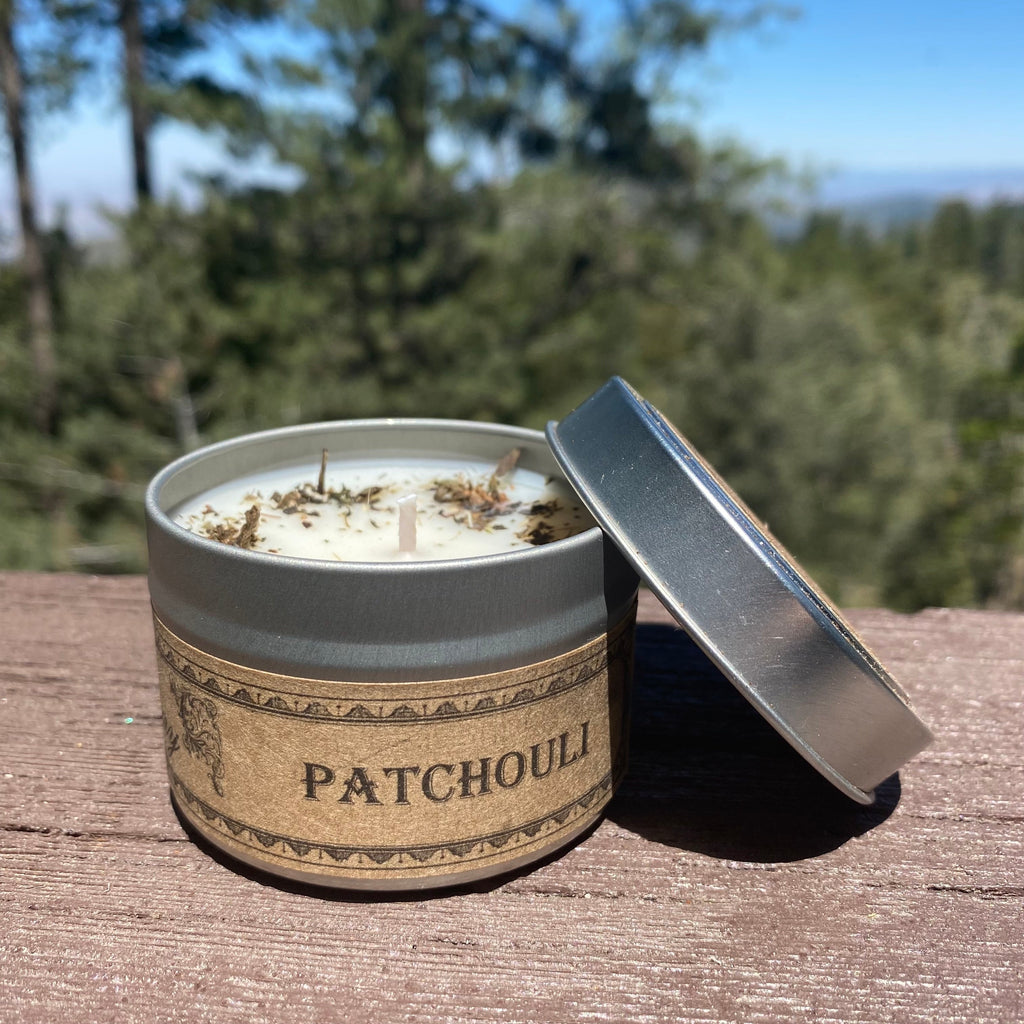 Candle in a 'Patchouli' scented tin with a natural label, outdoors on a wooden surface with trees in the background.