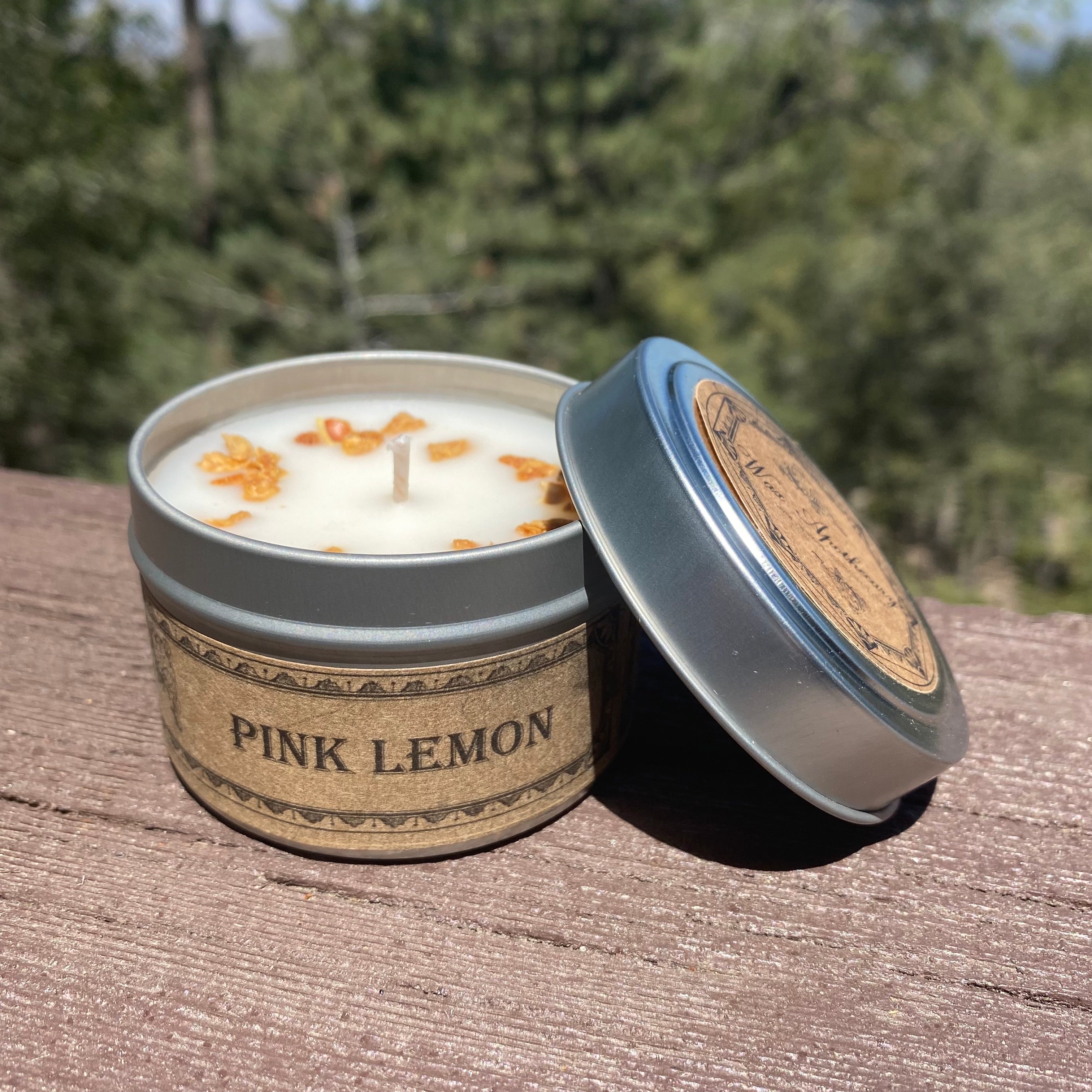 Candle in a tin labeled 'Pink Lemon' on a wooden surface with a blurred natural background