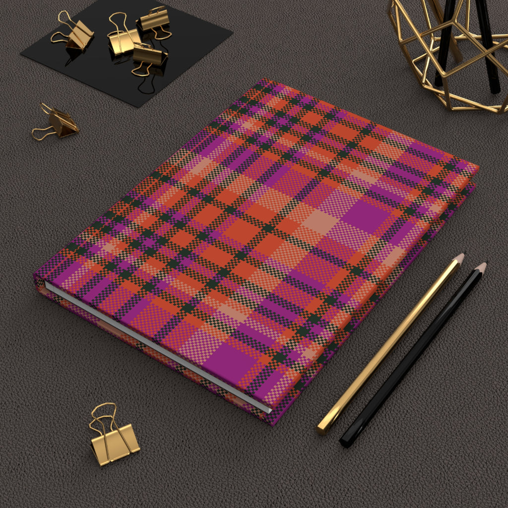 A hardcover notebook with a vibrant purple, orange, and pink plaid pattern, styled on a dark textured surface with gold and black office accessories.