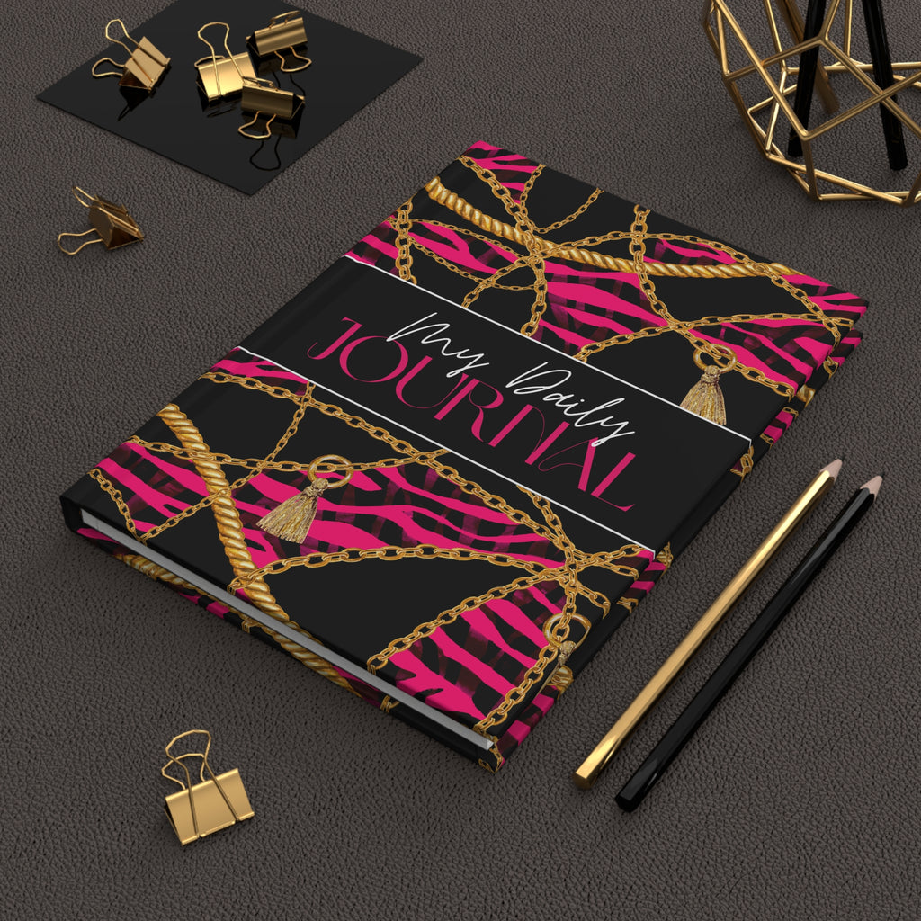 A hardcover journal with a black cover decorated with hot pink zebra stripes, gold chains, and tassels, styled on a dark textured surface with gold and black office accessories.